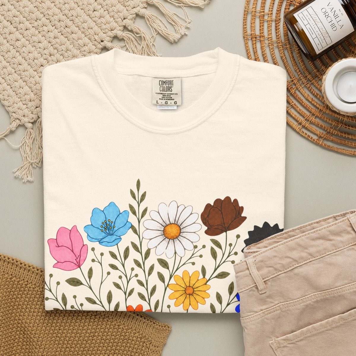 A folded off-white short-sleeve crewneck t-shirt featuring a graphic print of an open book with colorful LGBTQ colored wildflowers blooming from it, displayed on a clean white background