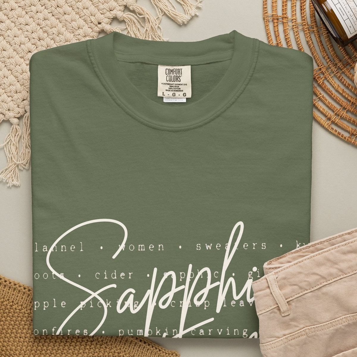 An olive-green crewneck t-shirt with a white script graphic reading 'Sapphic Fall' over a list of food types, styled in a flat lay with beige pants, a knit blanket, a candle jar, and a woven mat on a light gray background.