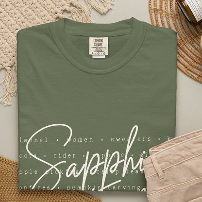 An olive-green crewneck t-shirt with a white script graphic reading 'Sapphic Fall' over a list of food types, styled in a flat lay with beige pants, a knit blanket, a candle jar, and a woven mat on a light gray background.