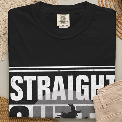A folded black graphic T-shirt is arranged in a styled flat lay with beige pants, a woven rug, a candle jar, and a small cup on a neutral surface. The shirt has a bold white and gray graphic across the chest reading 'STRAIGHT OUTTA THE BROOM CLOSET'