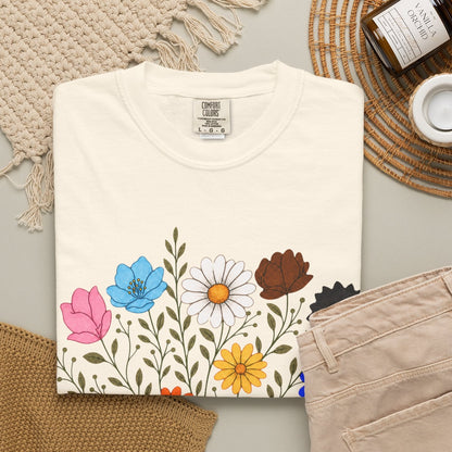 A folded off-white short-sleeve crewneck t-shirt featuring a graphic print of an open book with colorful LGBTQ colored wildflowers blooming from it, displayed on a clean white background