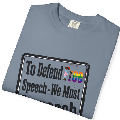 A folded slate blue t-shirt with a graphic print on the front. The graphic features a distressed sign design with the text 'To Defend Free Speech - We Must' in black, with 'Free' highlighted in a LGBTQ rainbow flag pattern.