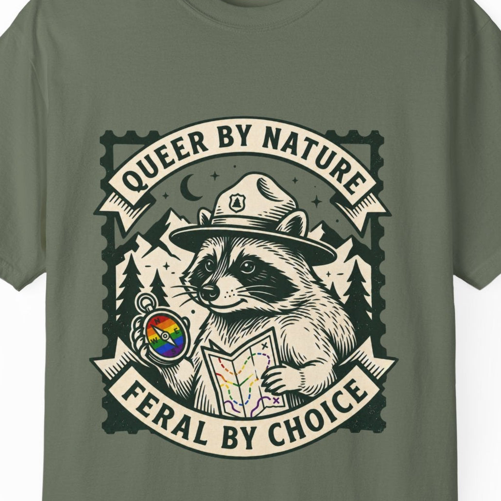 An olive green short-sleeve t-shirt featuring a large graphic print of the raccoon Jimothy dressed as a park ranger holding a map, with the text 'QUEER BY NATURE' and 'FERAL BY CHOICE'.