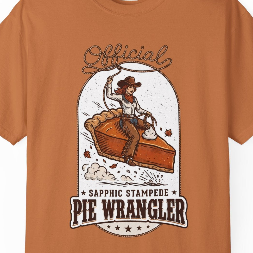 An orange short-sleeve crewneck t-shirt featuring a vintage-style graphic of a cowgirl riding a pie, with the text 'Official Sapphic Stampede Pie Wrangler' printed in brown and orange.