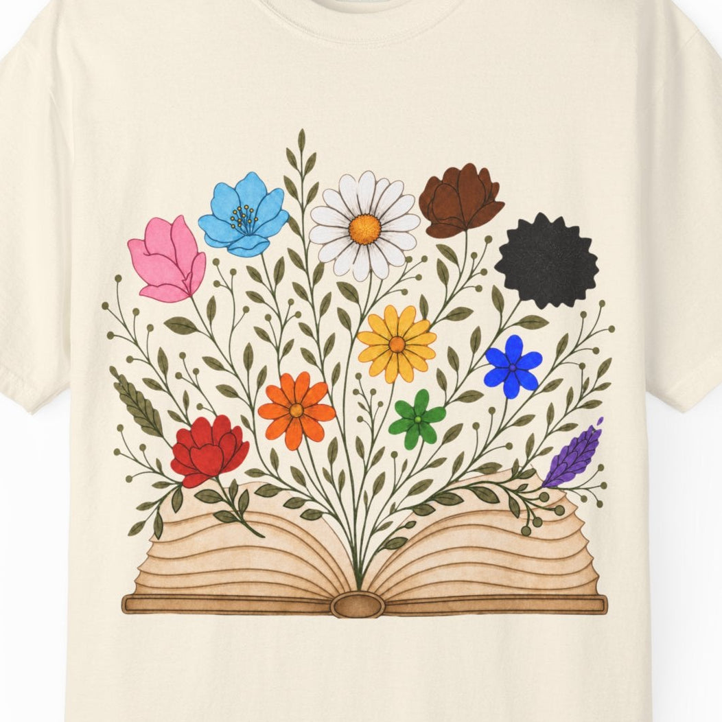 An off-white short-sleeve crewneck t-shirt featuring a graphic print of an open book with colorful LGBTQ colored wildflowers blooming from it, displayed on a clean white background