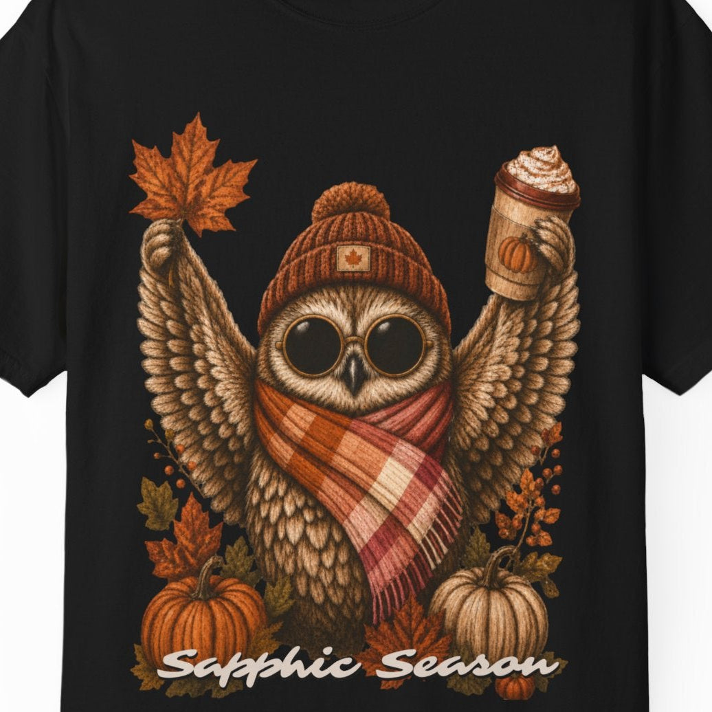 A black short-sleeve crewneck t-shirt featuring a detailed graphic of a cute owl wearing a knit beanie and a lesbian flag-colored plaid scarf, holding a hot drink. The graphic is decorated with autumn leaves, pumpkins, and the text 'Sapphic Season' at the bottom.