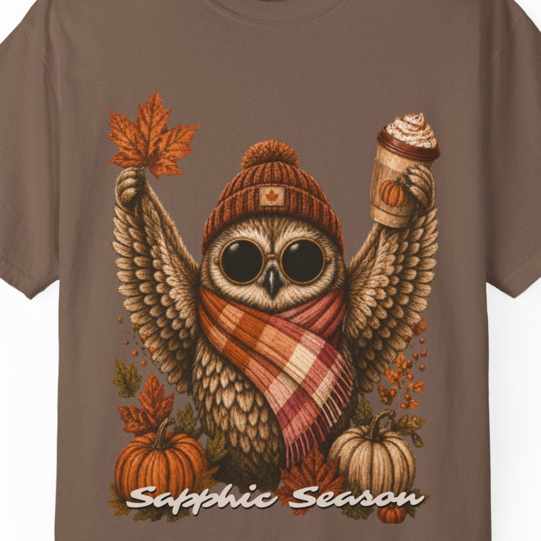 A brown short-sleeve crewneck t-shirt featuring a detailed graphic of a cute owl wearing a knit beanie and a lesbian flag-colored plaid scarf, holding a hot drink. The graphic is decorated with autumn leaves, pumpkins, and the text 'Sapphic Season' at the bottom.