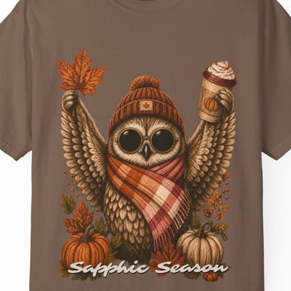 A brown short-sleeve crewneck t-shirt featuring a detailed graphic of a cute owl wearing a knit beanie and a lesbian flag-colored plaid scarf, holding a hot drink. The graphic is decorated with autumn leaves, pumpkins, and the text 'Sapphic Season' at the bottom.