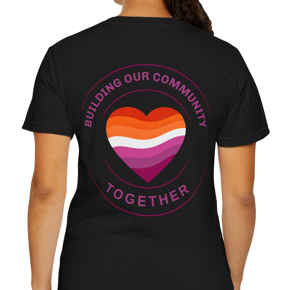 A woman shown from the back wearing a black short-sleeve t-shirt with a circular pride graphic on the back. The graphic features a heart divided into orange, white, pink, and purple stripes, with the words 'BUILDING OUR COMMUNITY TOGETHER' in pink around it.
