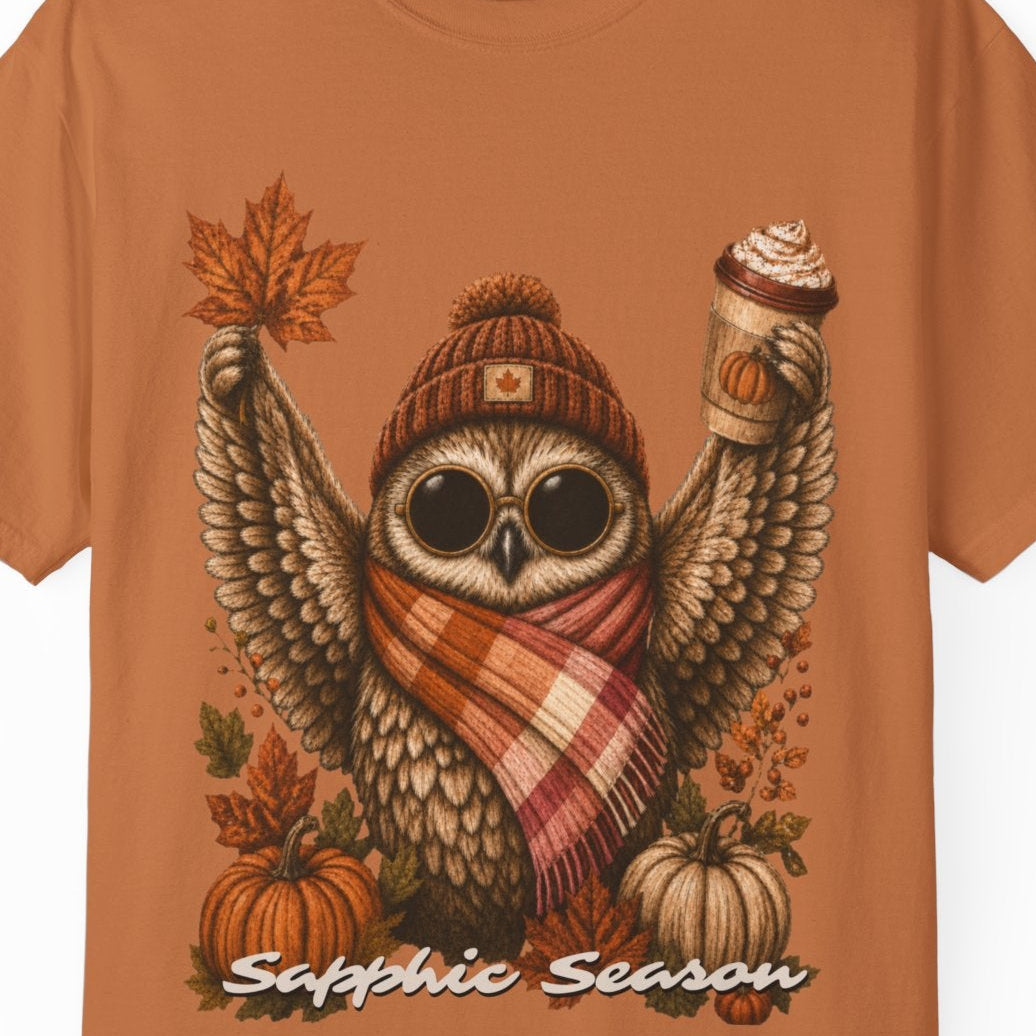 A rust colored short-sleeve crewneck t-shirt featuring a detailed graphic of a cute owl wearing a knit beanie and a lesbian flag-colored plaid scarf, holding a hot drink. The graphic is decorated with autumn leaves, pumpkins, and the text 'Sapphic Season' at the bottom.