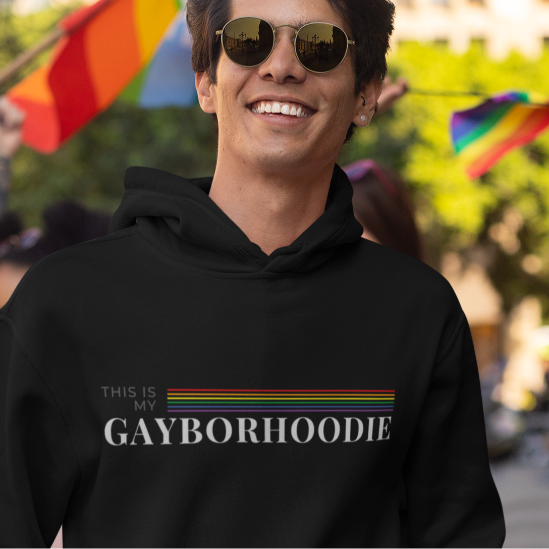 A mockup of a gay man wearing a LGBT Pride hoodie that is black in color for a Gay Pride tee that says "This is my GAYBORHOODIE" with LGBTIQQ rainbow colors| Pride sweatshirt designed by Queer Energy 