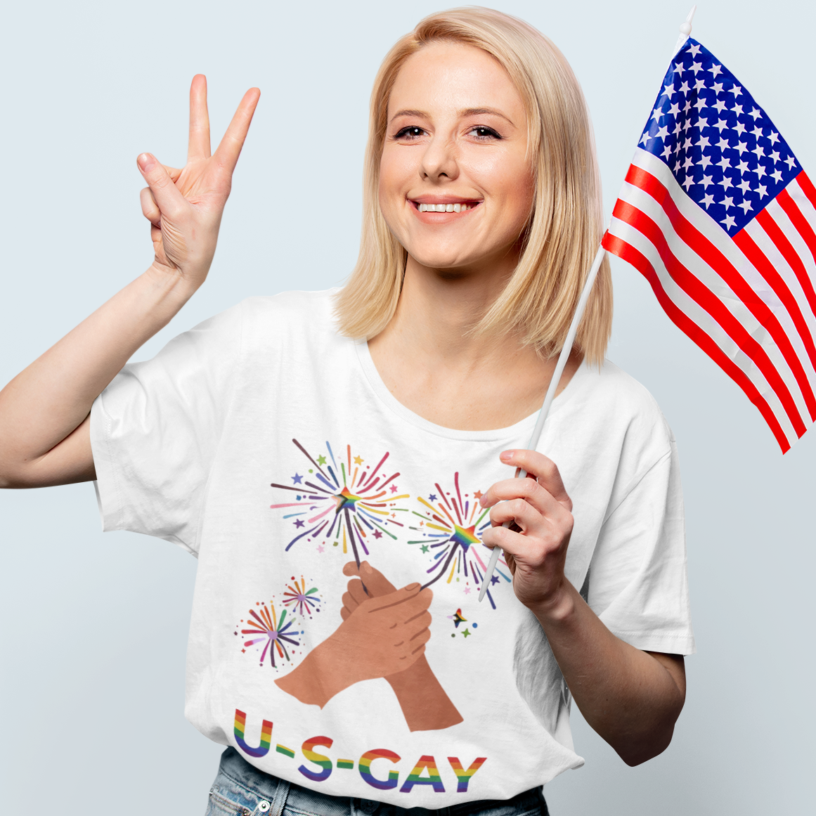 Lesbian wearing a Gay pride t-shirt featuring U.S. Gay rainbow text with colorful Pride fireworks, holding an American flag against a light gray background. Design by Queer Energy